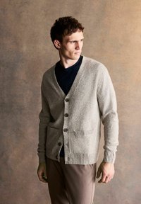 Light grey cardigan with a V-neck, button front, ribbed cuffs, and two front pockets, paired with a navy t-shirt and brown trousers.