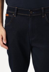 Jeans din denim albastru închis, cu cusături portocalii, design cu cinci buzunare și închidere cu nasture, având un eticheta de brand din piele pe talie.