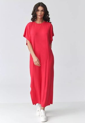 Kobieta z ciemnymi, kręconymi włosami, ubrana w luźną, czerwoną sukienkę maxi i białe trampki, stojąca na tle jednolitego, jasnoszarego tła.