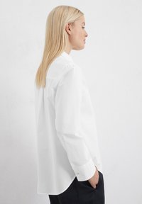 Camicia bianca con colletto a camicia e maniche lunghe, caratterizzata da un tessuto morbido e un orlo dritto. Colletto discreto e un unico bottone sui polsini.