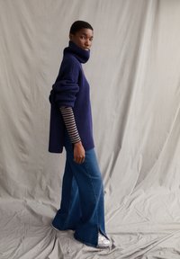 Überdimensionierter navy-blauer Ripp-Pullover mit hohem Kragen, über einem gestreiften Langarmshirt getragen, kombiniert mit blauen Schlagjeans und silbernen Schuhen.