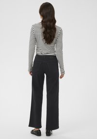 Femme aux longs cheveux ondulés portant un haut à rayures noires et blanches à manches longues, un pantalon large noir et des mocassins noirs, se tenant devant un fond uni.