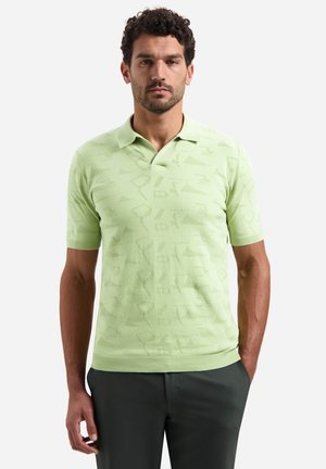 Man draagt een lichtgroene korte mouw gebreide polo met een subtiel geometrisch patroon, staand met handen in de zakken tegen een effen achtergrond.