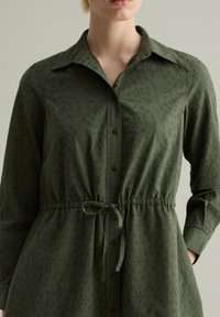 Olivgrünes Hemdkleid aus leichtem Material, mit durchgehender Knopfleiste, Kragen, langen Ärmeln und verstellbarem Taillenbund mit floraler Textur.