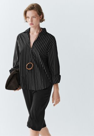 Kvinde iført en sort og grå skjorte med vertikale striber og opkalkede ærmer, sort nederdel, træringhalskæde og holder en mørk tekstureret clutch.