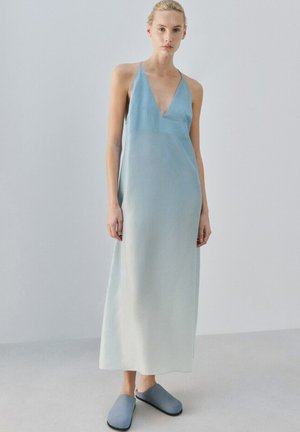 Kobieta o blond włosach nosi bezrękawnikową, maxi sukienkę w gradientowym odcieniu od niebieskiego do białego oraz niebieskie wsuwane buty na jasno szarym tle.