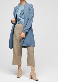 Cardigan lavorato a maglia blu chiaro con un motivo testurizzato, abbinato a una maglietta blu con una grafica a foglia e pantaloni larghi a quadri marroni.