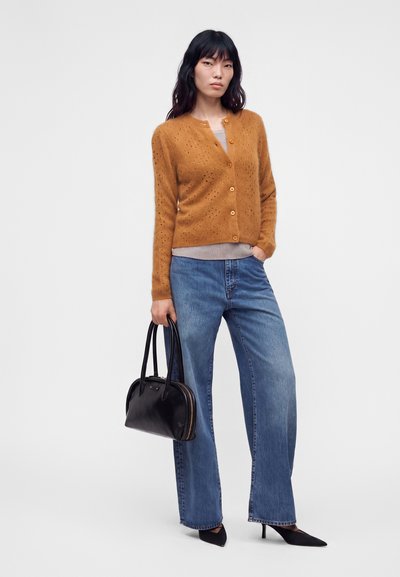 Femme portant un cardigan orange boutonné, un jean large bleu, des talons noirs pointus, tenant un sac à main noir sur un fond uni.