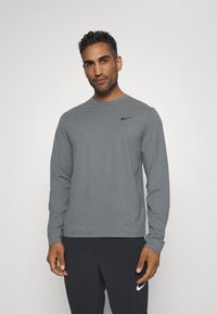 Hosszú ujjú szürke Nike póló, amely kerek nyakkivágással és egy kis fekete logóval rendelkezik a bal melle fölött. Simább, sportos szabású anyag.