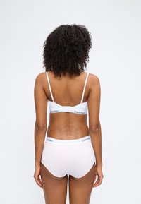 Γυναίκα με σγουρά μαλλιά που φοράει λευκό σουτιέν Calvin Klein και εσώρουχα με ψηλή μέση, απεικονιζόμενη από πίσω σε απλό φόντο.