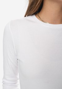 Weißes Langarmoberteil mit gerippter Textur, Rundhalsausschnitt und einem subtilen Schimmer durch eine goldene Kettenhalskette.