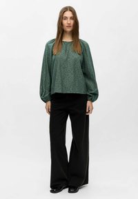 Vrouw met steil bruin haar in een groene blouse met lange mouwen en brede zwarte broek, staand voor een eenvoudige witte achtergrond.