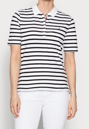 Femme portant un polo blanc à rayures horizontales noires et un pantalon blanc, debout devant un fond clair uni.