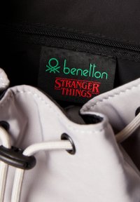 Szara materiałowa torba z białymi sznureczkami, z czarną metką z napisem "benetton" na zielono i "STRANGER THINGS" na czerwono. Zamykana na zamek.