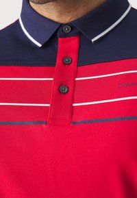 Červená polo košile s námořnickým límcem, s bílými a tmavě modrými vodorovnými pruhy. Pět tlačítek s černými knoflíky. Logo Calvin Klein na boku.