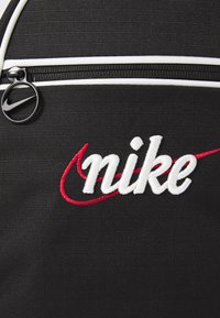 Rucsac negru din țesătură texturată, cu fermoar alb și un snur circular negru pentru fermoar. Prezintă un logo brodat "nike" în alb și roșu.