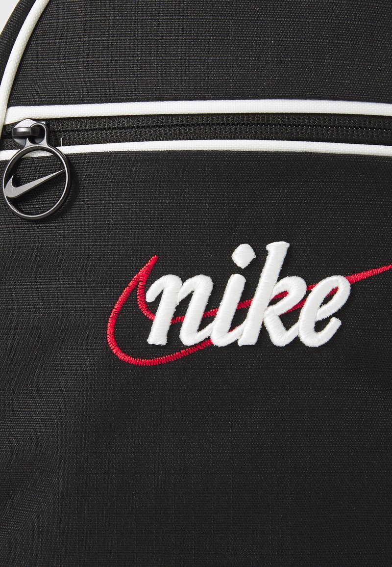 Czarny plecak z teksturowanej tkaniny, z białym zamkiem błyskawicznym i okrągłym czarnym uchwytem zamka. Posiada haftowane logo "nike" w kolorze białym i czerwonym.