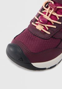 Καφέ μπορντό παπούτσι πεζοπορίας με υφασμάτινο άνω μέρος με υφή, μαύρο λαστιχένιο κάλυμμα στα δάχτυλα και ελαστικά κορδόνια. Διαθέτει ενισχυμένη μεσαία σόλα για υποστήριξη.