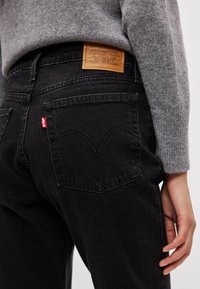 Persona, kas valkā melnas Levi's džinsas un pelēku džemperi, rādot aizmugurējās kabatas detaļas un ādas zīmola uzlīmi pie jostasvietas.