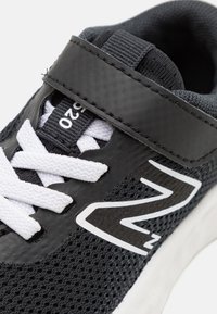 Baskets de sport noires avec une tige en mesh, lacets blancs et une sangle. Dotées d'un logo bien visible et d'une semelle en caoutchouc texturé pour une bonne adhérence.