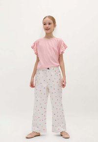 Helles rosa T-Shirt mit Rüschensleeves, kombiniert mit weißen, weit geschnittenen Hosen mit floralem Muster in Orange und Lila. Einfache Ballettschuhe.