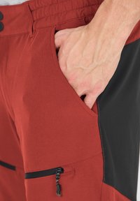 Röda byxor med svarta detaljer har en säker ficka med dragkedja och en sidoficka för handen, tillverkade av ett slätt, strukturerat tyg. Elastisk midja synlig.