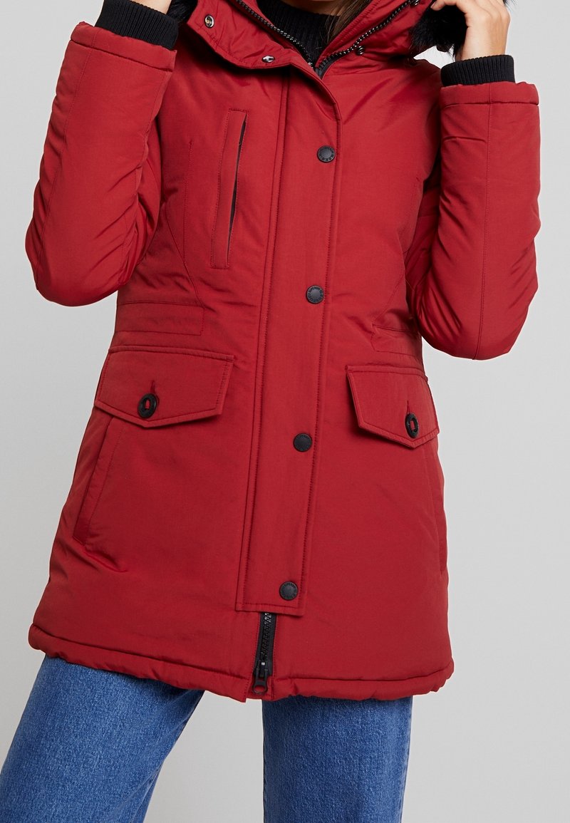 Veste d'hiver isolante rouge avec boutons noirs, fermeture éclair, poche poitrine, deux poches à rabat et capuche bordée de fausse fourrure, portée avec un jean bleu.