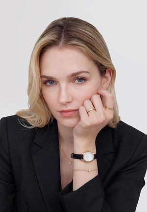 Vaaleahiuksinen nainen mustassa bleiserissä, joka nojaa leukaa käteensä ja käyttää kultasormuksia, mustaa kelloa ja hienoja kultaisia rannekoruja.