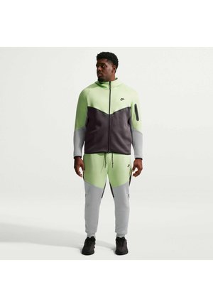 Mand iført grøn, sort og grå Nike træningsdragt med hættejakke og joggingbukser, stående mod en ensartet lys baggrund.