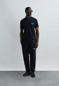 Άνδρας που φοράει μαύρο πόλο Moschino, ταιριαστό παντελόνι, μαύρα αθλητικά παπούτσια και γυαλιά ηλίου, στέκεται μπροστά σε ένα απλό ανοιχτό γκρι φόντο.