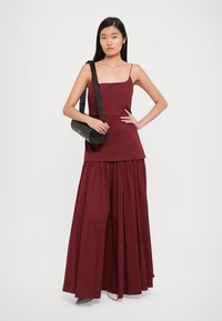 Burgundy sleeveless top paired with a flowy, pleated maxi skirt. Model holds a black textured handbag with a shoulder strap.