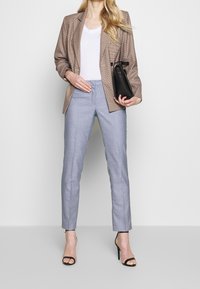Bruin geruit blazer over een witte top, gecombineerd met lichtblauwe tailleurbroek. Model houdt een zwarte handtas vast en draagt zwarte sandaaltjes met banden.