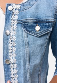 Chaqueta de mezclilla en azul claro, con botones de metal plateado y un borde de encaje blanco en la parte delantera, con un pequeño bolsillo en el pecho y un diseño clásico.