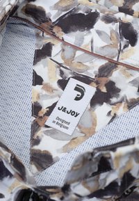 Stoffen met bloemenpatroon in beige, bruin en zwart; blauwe streepvoering; wit label met "J&JOY" en "Ontworpen in België."