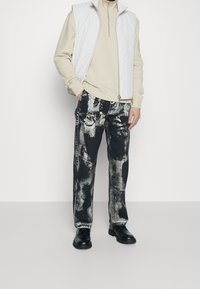 Chaleco blanco sin mangas sobre un cuello de tortuga beige, combinado con jeans negros que tienen un patrón desgastado en blanco y gris, y zapatos negros.