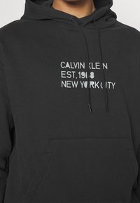 Felpa con cappuccio nera in cotone con tasca frontale. Presenta un testo riflettente: "CALVIN KLEIN EST.1968 NEW YORK CITY." Design minimalista.