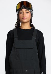 Zwarte sneeuwbib met verstelbare banden, grote voorklep en textuurstof. Draag over een zwart longsleeve shirt. Inclusief reflecterende accenten.