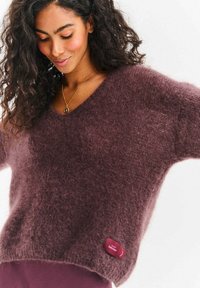 Przewymiarowany sweter z głębokim dekoltem w serek w kolorze bordo, wykonany z miękkiego, puszystego materiału. Posiada małą naszywkę z logo na dole.