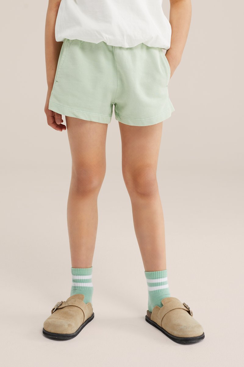 Enfant portant un short vert clair, des chaussettes rayées vertes et blanches assorties, et des chaussures beige à enfiler, debout devant un fond uni.