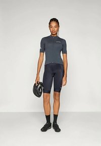 Cyklistická dres s krátkym rukávom v tmavosivej farbe, priliehavý strih s predným zipsom, v kombinácii s čiernymi šortkami a cyklistickými topánkami. Drží čiernu prilbu.