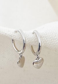 Los pendientes de aro de plata presentan pequeños charms en forma de corazón que cuelgan. El acabado liso refleja la luz, destacando su superficie pulida y diseño minimalista.