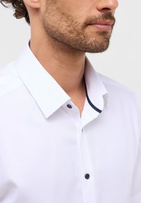 Camicia bianca con colletto aperto, dettagli dei bottoni scuri e un motivo a pois all'interno del colletto. Tessuto liscio.