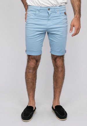 Homme portant un short bleu clair retroussé, une chemise blanche et des chaussures noires sans lacets, debout sur un fond gris uni.