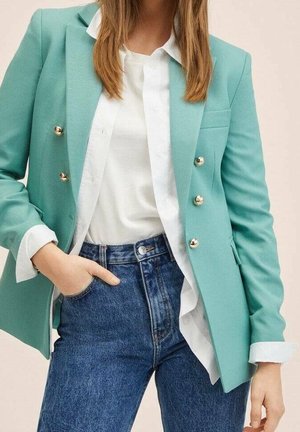 Vrouw draagt een muntgroene double-breasted blazer, wit bloesje met knopen en blauwe high-waisted jeans met één hand in de zak.