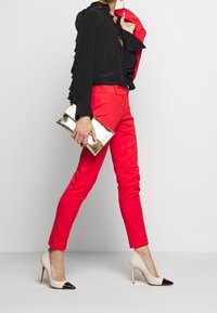 Blouse noire à volants, pantalons rouges ajustés, talons blancs avec embouts noirs, pochette dorée. Modèle marchant sur fond uni.