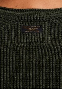Texture di un maglione lavorato a maglia verde scuro con un motivo a costine. L'etichetta recita: "LIBERTÀ ILLIMITATA, TESTATO AD ALTE ALTITUDINI, TIPO N° C-46."