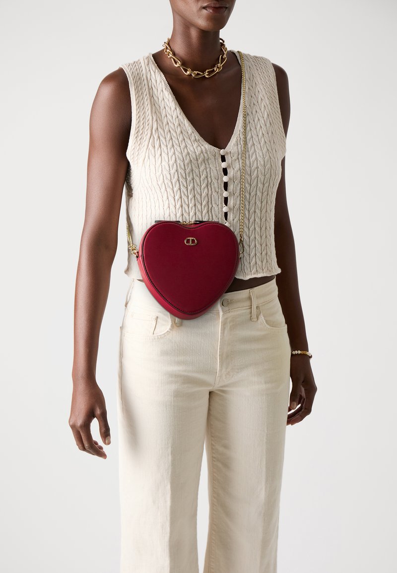 Woman wearing sleeveless beige knit vest and cream pants, accessorized with chunky gold necklace, bracelet, and red heart-shaped crossbody bag.