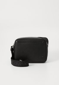 Black textured leather crossbody bag with a wide Calvin Klein branded fabric strap and small embossed cK logo on front.