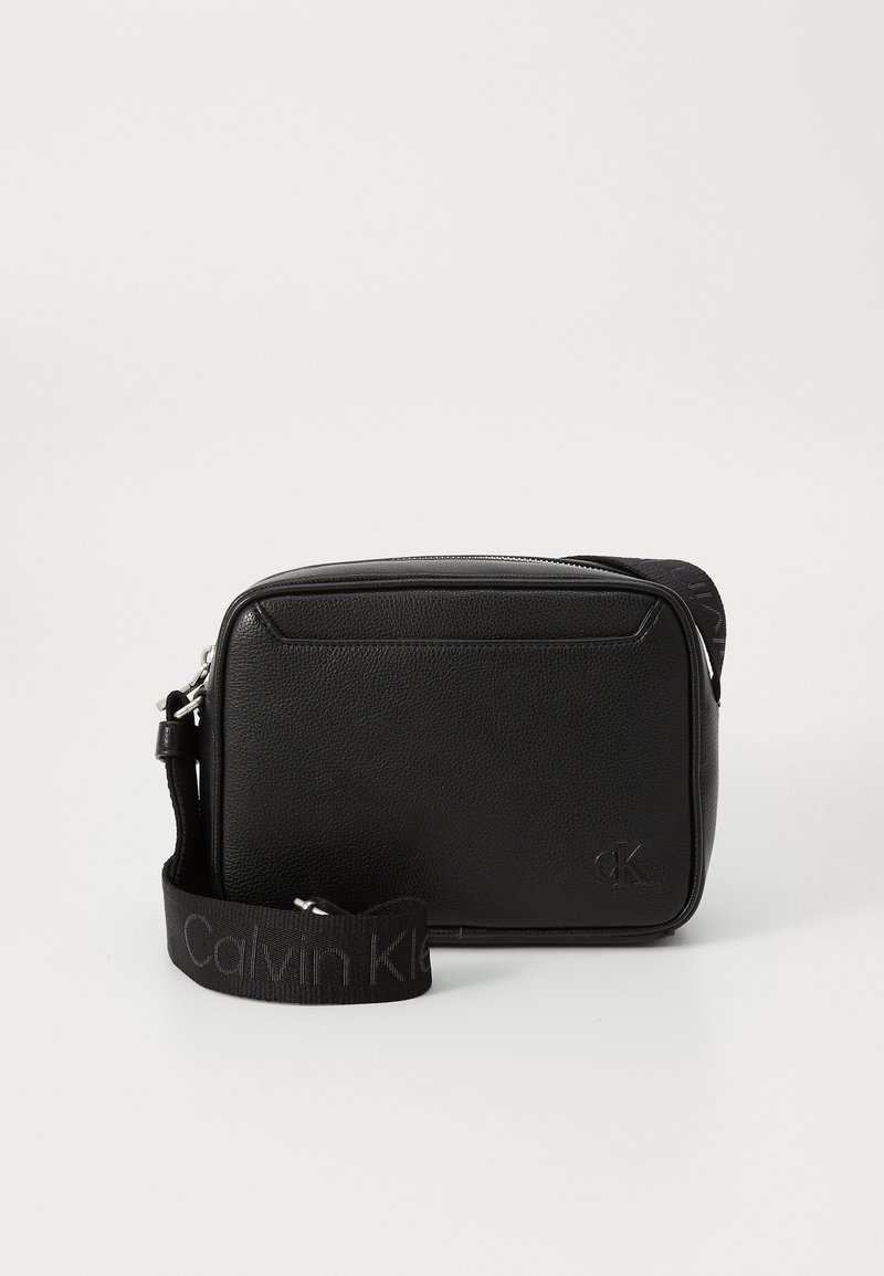 Black textured leather crossbody bag with a wide Calvin Klein branded fabric strap and small embossed cK logo on front.