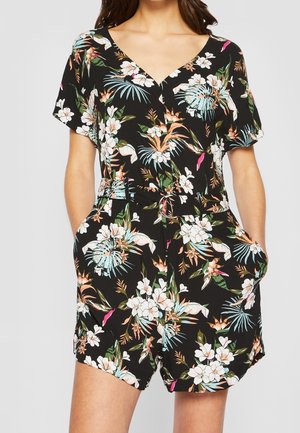 Woman wearing black short-sleeve floral romper with V-neck, waist tie, and side pockets, featuring white, green, blue, and peach tropical flowers.